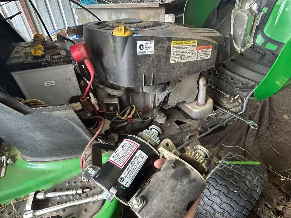 Replacement starter motor held beside a ride on mower engine in a shed